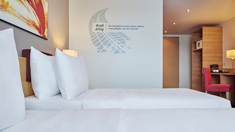 A modern hotel room with two comfortable beds and light decor. On the wall, there is a decorative graphic as well as a desk in the background.