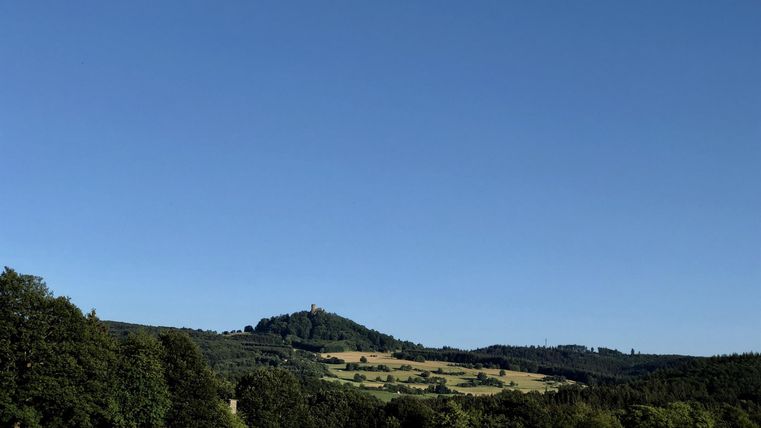 Eine ruhige Landschaft mit sanften Hügeln und einem klaren blauen Himmel. Im Vordergrund sind grüne Wiesen und Bäume zu sehen.