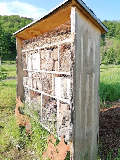Ein Insektenhotel aus Holz mit verschiedenen Materialien für Nützlinge steht in einer grünen Wiese. Im Vordergrund sind Tier-Silhouetten sichtbar.