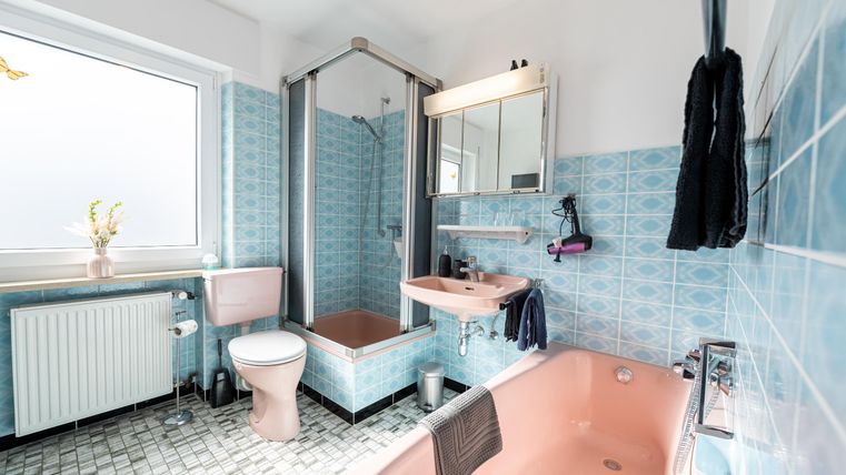 Een lichte badkamer met blauwe tegels en een badkuip. Er is een douche, een wastafel en een raam dat natuurlijk licht binnenlaat.