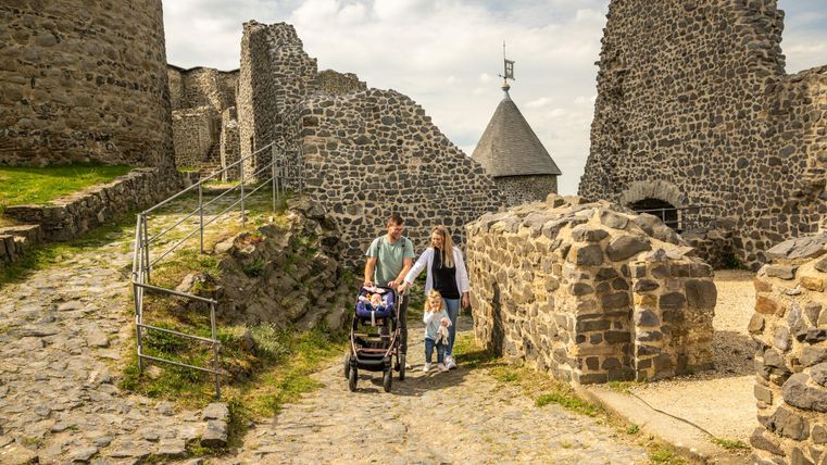 Eine Familie mit einem Kinderwagen besucht die Überreste einer alten Burg. Der Weg ist von Steinen und Gras umgeben, mit alten Mauern im Hintergrund.