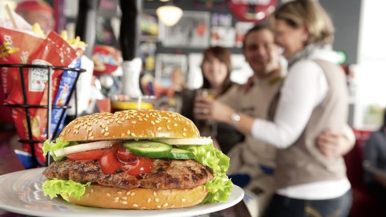 Un burger juteux avec un pain au sésame, de la laitue et des légumes frais. À l'arrière-plan, des invités joyeux sont visibles dans un environnement animé.