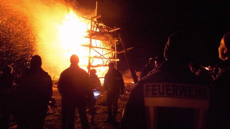 Ein großes Feuer brennt nachts an einem Holzgerüst. Mehrere Personen stehen in sicherer Entfernung und beobachten die Flammen.