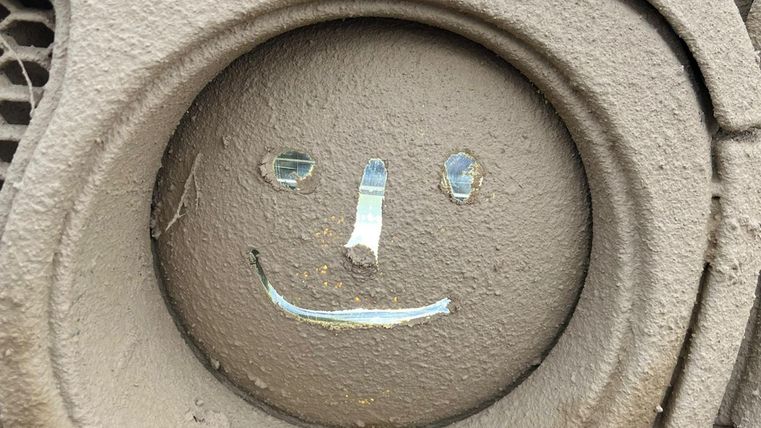 Ein lächelndes Gesicht aus grauem Material, das aus einer runden Oberfläche gemacht ist. Die Augen und der Mund sind mit kleinen, glänzenden Details hervorgehoben.