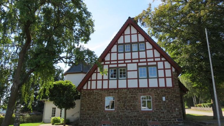 Ein traditionelles Fachwerkhaus mit einem roten Dach und großen Fenstern. Umgeben von grünem Gras und Bäumen.