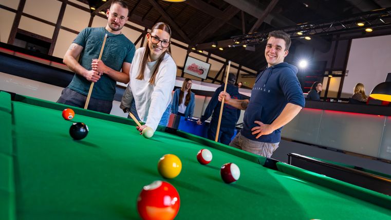 Three young people are standing at the pool table and smiling. Colorful pool balls are lying on the table.