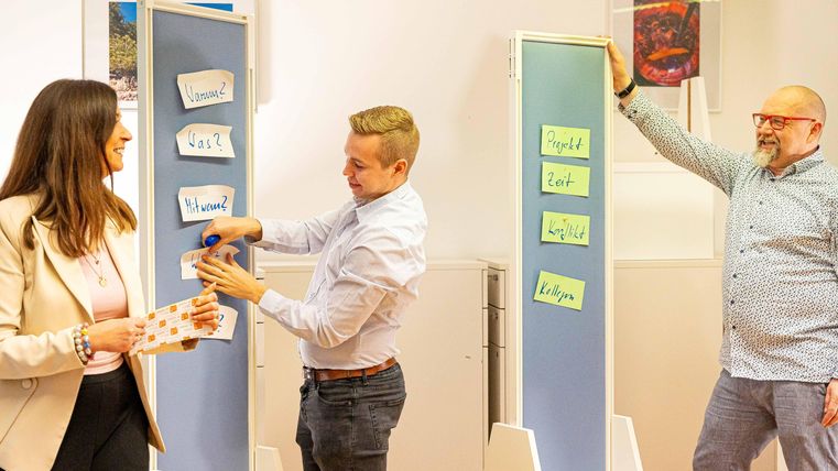 Drei Personen arbeiten zusammen in einem modernen Büroraum. Sie nutzen Pinnwände mit Notizen, um Ideen auszutauschen und zu diskutieren.