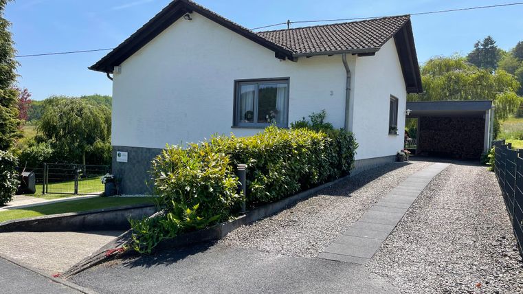 Une maison confortable avec un jardin bien entretenu et une allée. Le ciel est clair et bleu, entouré de paysages verdoyants.