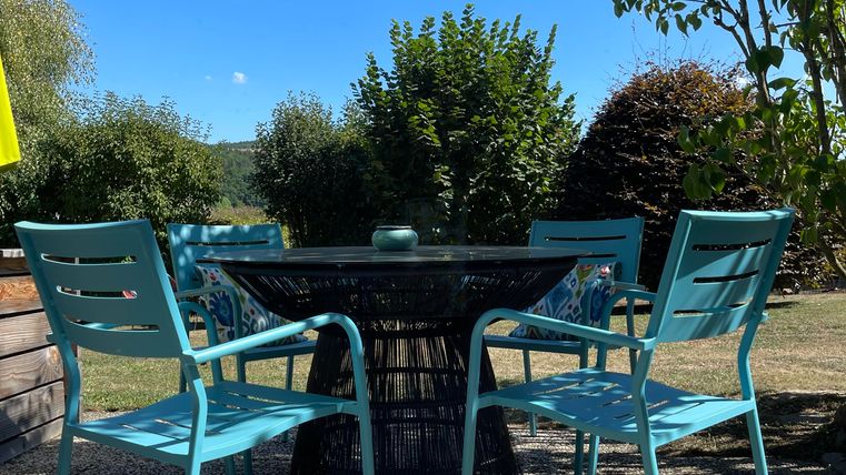 Une table avec quatre chaises bleues à l'extérieur. En arrière-plan, on peut voir des arbres et un ciel bleu clair.