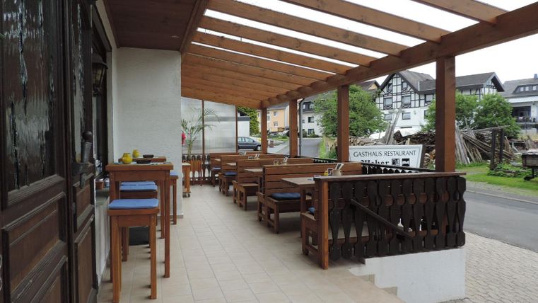 Ein restaurantähnlicher Außenbereich mit hölzernen Tischen und Stühlen unter einem überdachten Vordach. Die Umgebung ist grün und ruhig.