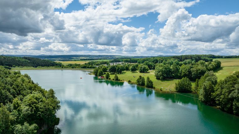 Ein ruhiger See umgeben von grünen Wäldern und Wiesen. Der Himmel ist klar mit großen, weißen Wolken.