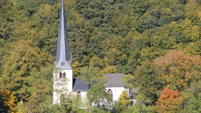 Kapelle mit spitzem Turm vor herbstlichem Wald.