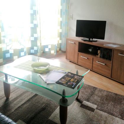 Ein modernes Wohnzimmer mit einem Glastisch und einem Fernseher. Die bunten Vorhänge lassen viel Licht herein.