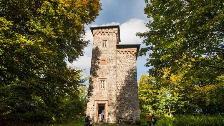 Ein historischer Steinturm umgeben von üppigem Grün und buntem Laub. Der Himmel ist teilweise bewölkt und sorgt für eine stimmungsvolle Atmosphäre.