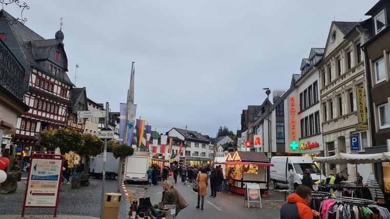 Ein belebter Marktplatz mit bunten Ständen und Menschen. Die Atmosphäre ist lebhaft und freundlich, umgeben von historischen Gebäuden.
