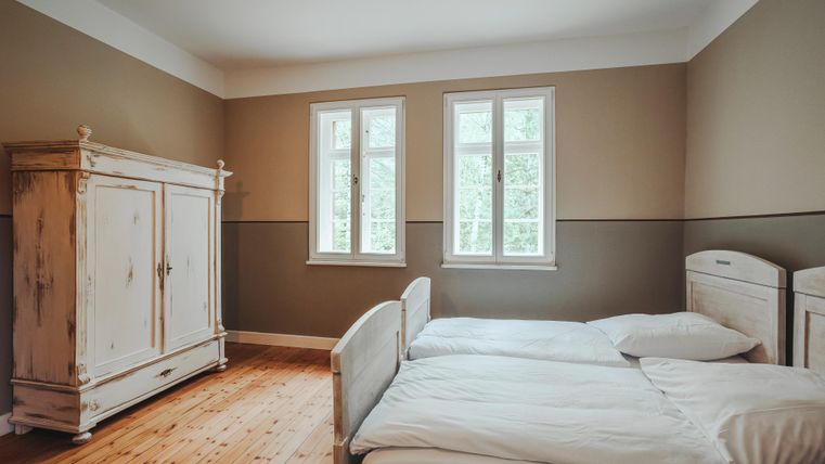 This image shows a bedroom with two single beds and a large antique wardrobe. Through two windows, one has a view of the trees outside.