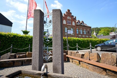 Ein moderner Brunnen aus Stein steht im Vordergrund, umgeben von Bänken und einer grünen Hecke. Im Hintergrund ist ein historisches Gebäude mit roten Ziegeln und Fahnen zu sehen.