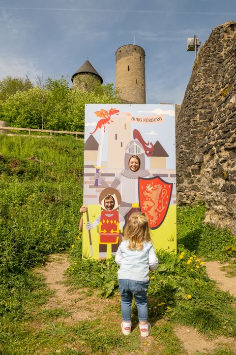 Vor der Burg Nürburg steht eine bunte Fototafel mit Rittermotiv und Ausschnitt für das Gesicht. Ein Kind blickt von hinten zur Tafel, während im Hintergrund die Burgmauern und Türme über grünem Bewuchs aufragen.