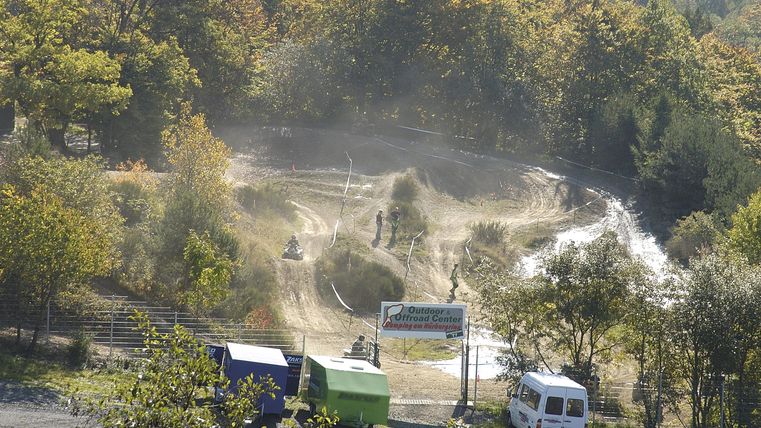 An off-road track with a dusty terrain and some vehicles. Surrounded by trees and natural vegetation.