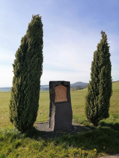 Ein Gedenkstein umgeben von zwei Zypressen. Die Landschaft ist grün mit einem klaren Himmel im Hintergrund.