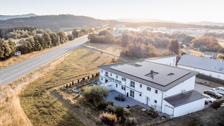 Een modern gebouw in een landelijke omgeving, omringd door weilanden en bomen. Op de voorgrond loopt een weg die in de verte leidt.
