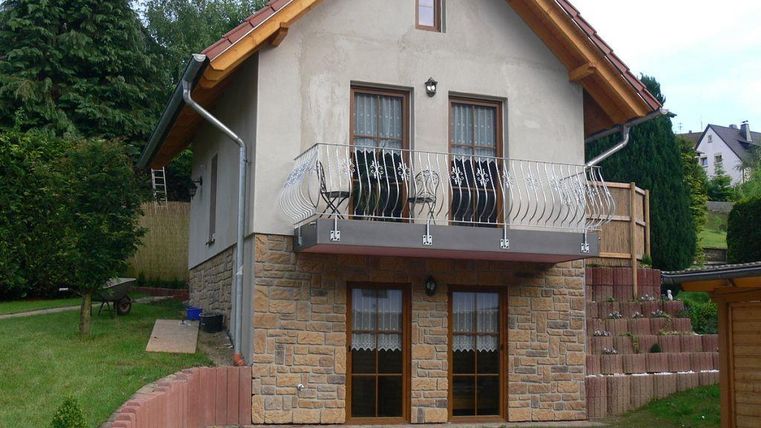 Een gezellig huis met een balkon en een stenen fundering. De tuin is onderhouden en omgeven door bomen.