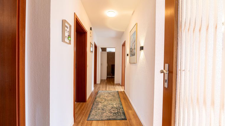 Een lichte gang met houten vloeren en witte muren. Aan de muren hangen schilderijen, en een tapijt geeft de ruimte een gezellige sfeer.