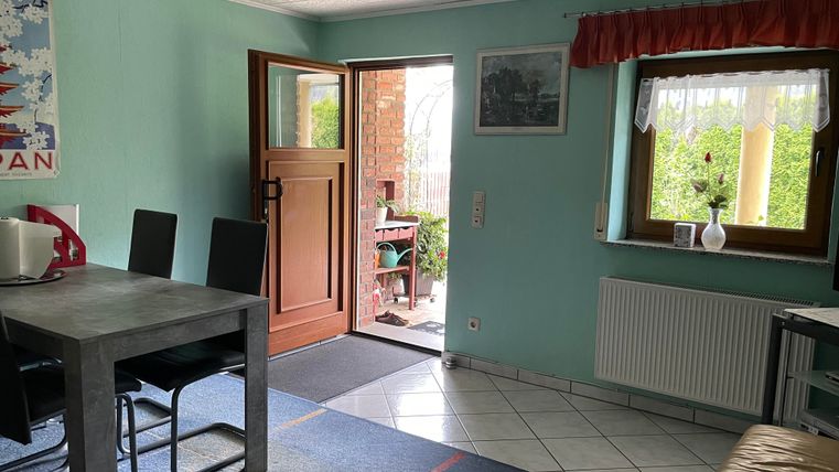 A cozy entrance area with a table and chairs. The door leads to a garden area with plenty of green plants.