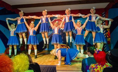 Een groep danseressen in blauwe kostuums voert een optreden uit. Op de achtergrond zijn kleurrijke clowns te zien.
