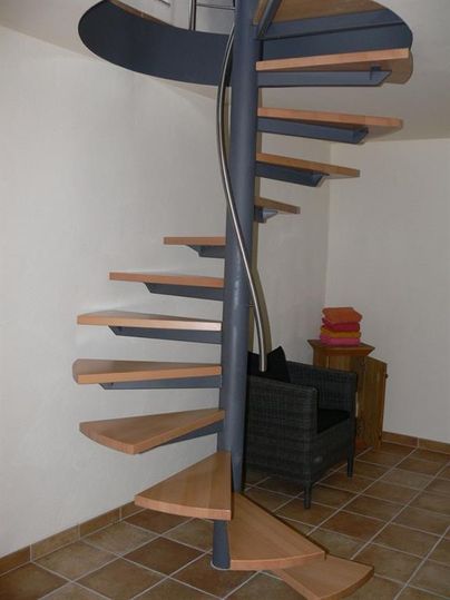 Een moderne draaitrap met houten treden en een stabiele metalen constructie. Naast de trap staat een comfortabele stoel en een kleine tafel.