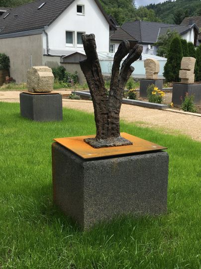 Eine moderne Skulptur aus Metall steht auf einem Steinsockel. Sie ist von einem grünen Rasen und verschiedenen anderen Kunstwerken umgeben.