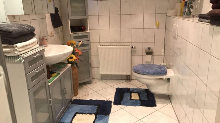 A modern bathroom with white tiles and a washbasin. There is a toilet area and decorations like towels and a small bouquet.