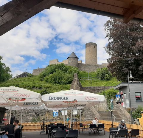 An old castle rises on a hill, surrounded by green trees and blue sky. In the foreground, there are tables and chairs of a café with sun umbrellas.