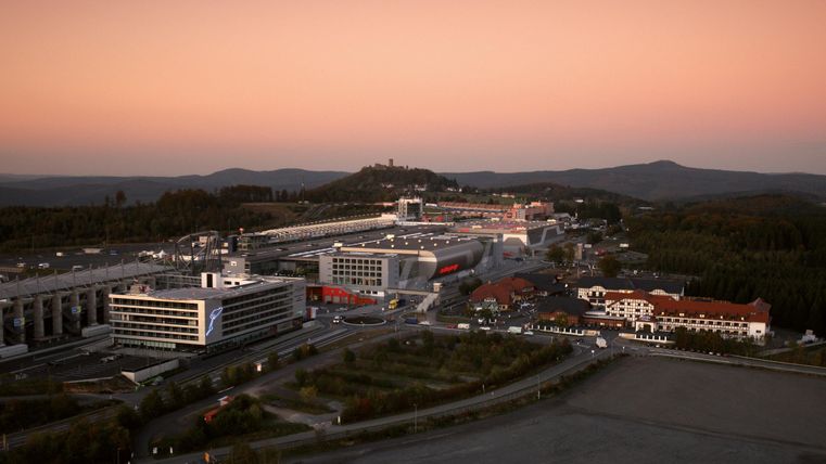 An industrial landscape with modern buildings and roads. The sky is colored in soft shades of pink and blue, while hills are silhouetted in the background.