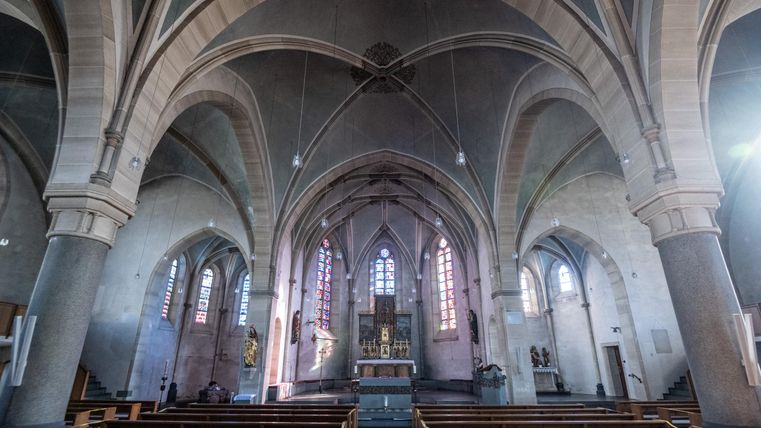 Ein großer, heller Kirchenraum mit hohen, gewölbten Decken und bunten Fenstern. Bänke sind auf beiden Seiten des Raumes angeordnet, während der Altar im Vordergrund steht.