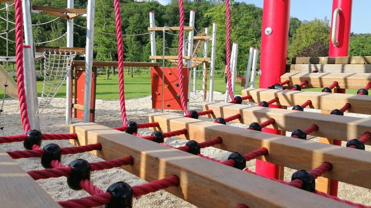 Ein Spielplatz mit Holzstrukturen und roten Seilen. Im Hintergrund sind grüne Bäume und ein klarer Himmel zu sehen.