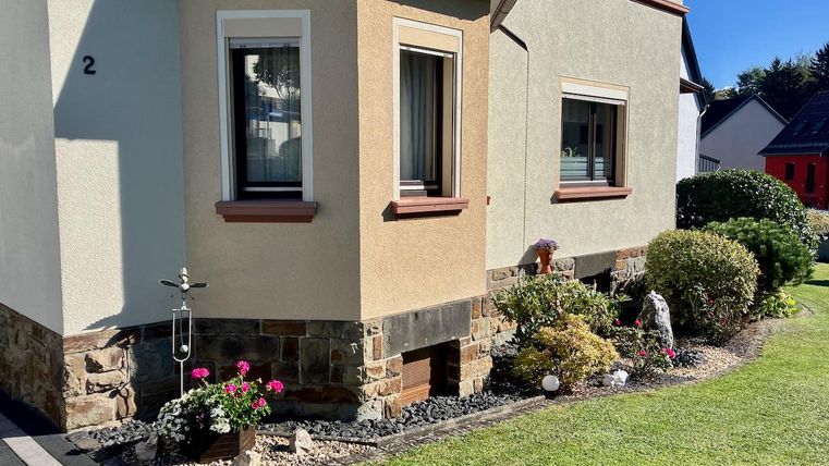 Une maison moderne avec un crépi extérieur lumineux et des fenêtres brunes. Le jardin est soigné, avec des fleurs colorées et des buissons.