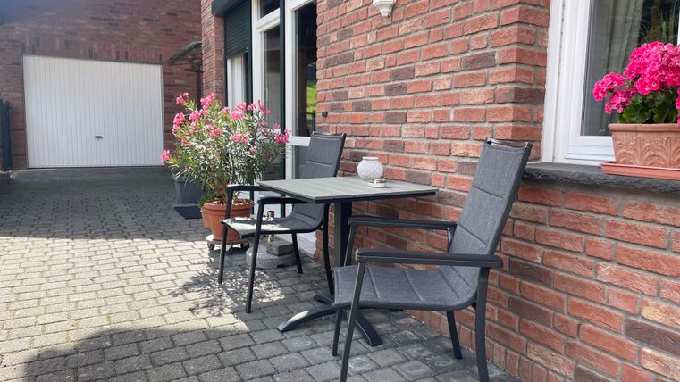 Een gezellige buitenruimte met een tafel en twee stoelen. Bloemen en een smaakvolle stenen muur omringen de plaats.