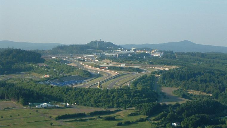 Un vaste paysage avec une piste de course et des collines douces à l'arrière-plan. Il y a des champs verts et quelques bâtiments à proximité de la piste.