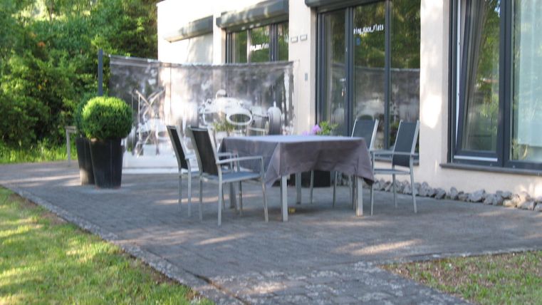 Ein gemütlicher Außenbereich mit einem Tisch und Stühlen auf einer Terrasse. Im Hintergrund sind große Fenster und ein gepflegter Garten zu sehen.