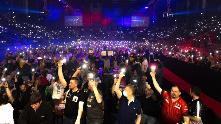 Eine große Menschenmenge in einer Arena, die mit ihren Handys Lichter zeigt. Die Atmosphäre ist feierlich und bunt beleuchtet.