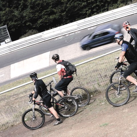 Vier Mountainbiker stehen an einem Zaun und beobachten ein Auto, das auf einer Rennstrecke vorbeif&auml;hrt., &copy; Markus Risse