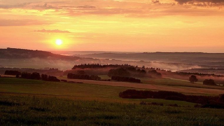 A beautiful sunset over gentle hills. The landscape is green with some trees in the foreground.