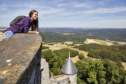 Eine Frau schaut über die Mauer einer Burg auf die umliegende Landschaft. Im Hintergrund sind grüne Hügel und ein blauer Himmel zu sehen.