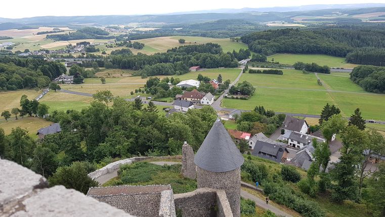Eine Aussicht von einer Burg über grüne Felder und kleine Dörfer. Die Landschaft ist hügelig mit vielen Bäumen und einem bewölkten Himmel.