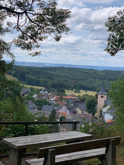 Ein schöner Ausblick auf ein kleines Dorf mit einem Kirchturm und umgebenden Wäldern. Der Himmel ist teilweise bewölkt und die Landschaft wirkt friedlich.