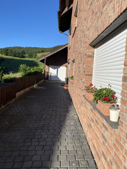 Een geplaveide weg loopt langs een bakstenen huis met bloeiende planten op de vensterbanken. De zonnige lucht en het groene landschap op de achtergrond creëren een uitnodigende sfeer.