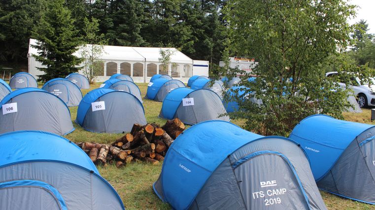 A campsite with several blue tents and a pile of wood in the foreground. In the background, there is a large tent.