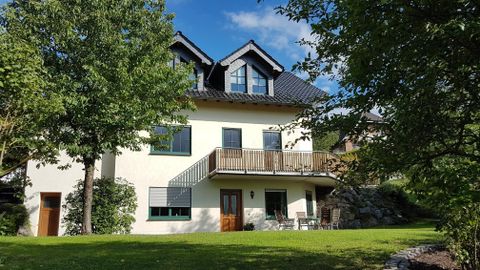 Ein modernes Haus mit einem Balkon und einem gepflegten Garten. Umgeben von Bäumen und unter einem klaren Himmel.