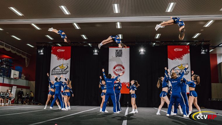 Een groep cheerleaders voert acrobatische stunts uit op een podium. Sommige deelnemers vliegen door de lucht, terwijl anderen de grond verlaten.